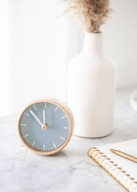 An analog clock on top of a marble table with a white vase in the back