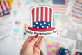 Load image into Gallery viewer, Hand holding a 'Happy Constitution Day' crown with an American flag design against a blurred background of flags.
