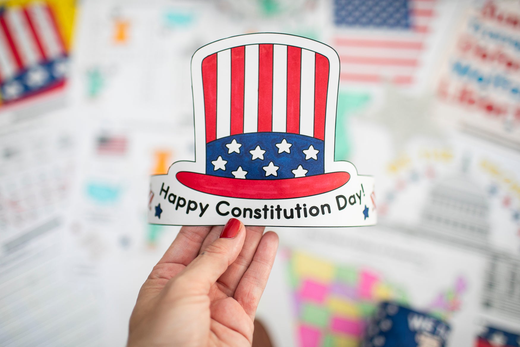 Hand holding a 'Happy Constitution Day' crown with an American flag design against a blurred background of flags.